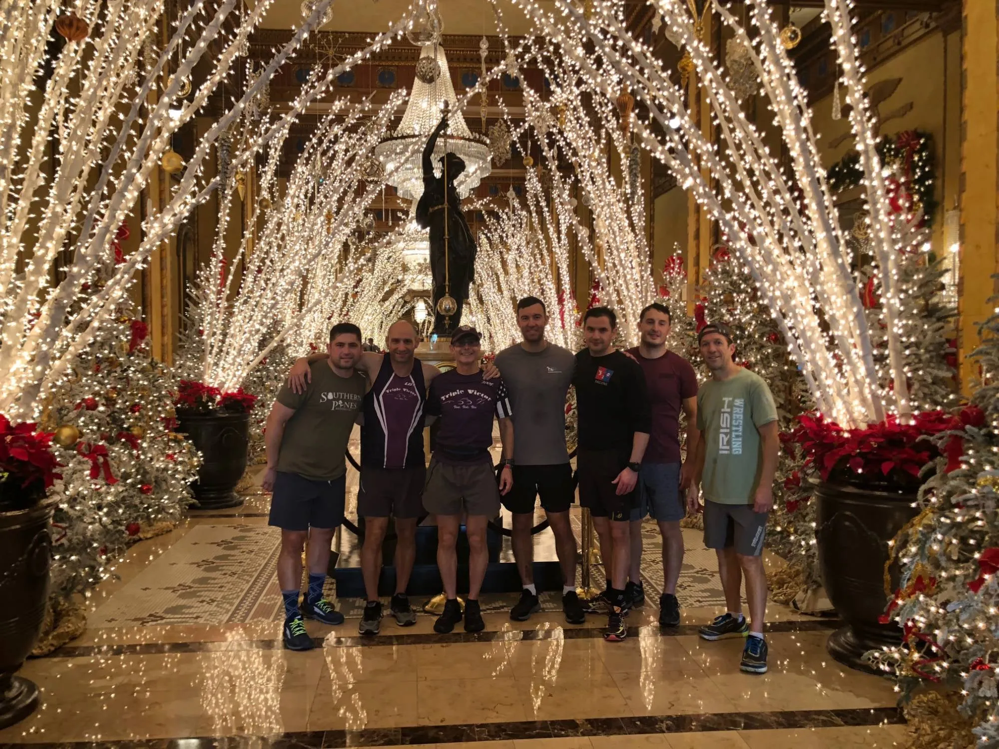 Dave and six friends in running clothes in The Roosevelt's lobby, under arches of white lights, with Christmas trees and red poinsettias on either side